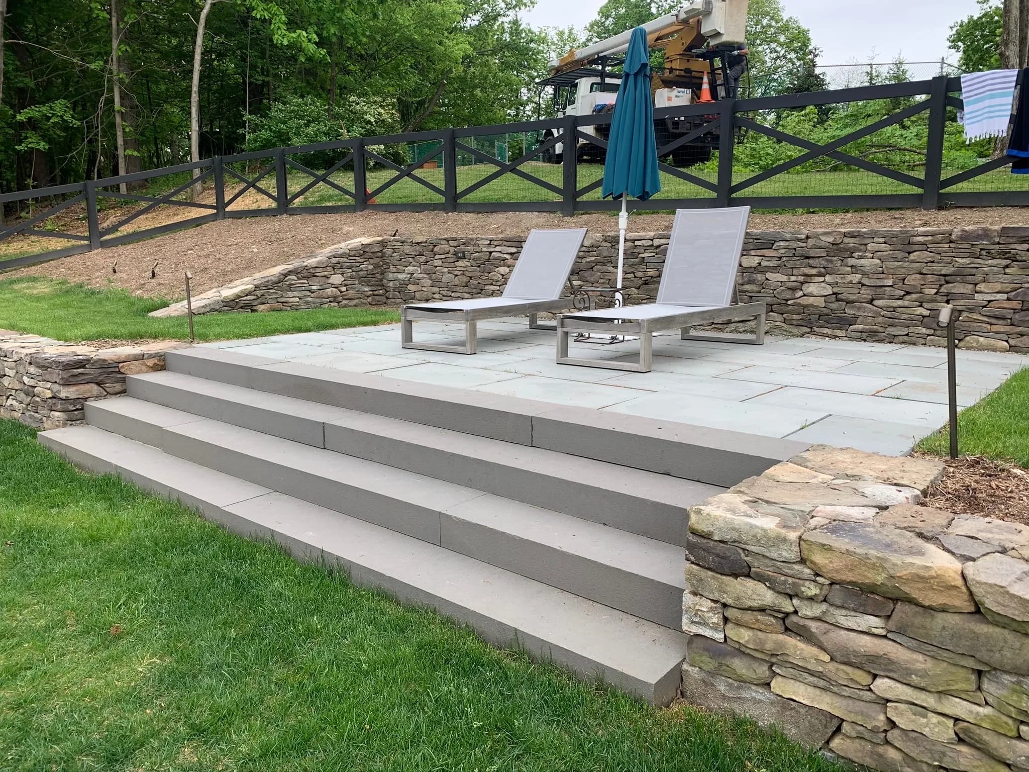 Outdoor patio with stone steps, lounge chairs, and an umbrella surrounded by greenery.